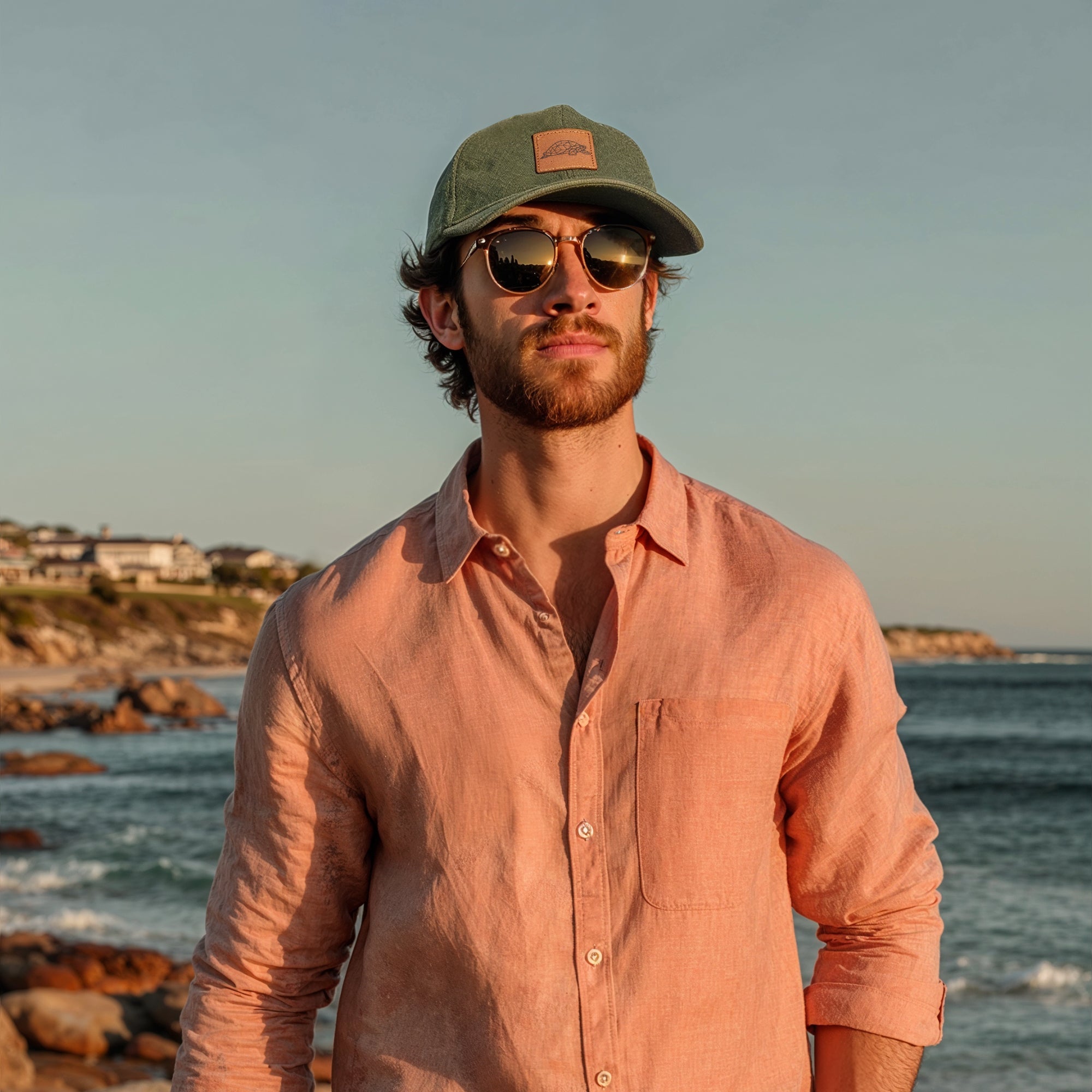 Man wearing a green cap and sunglasses standing by the ocean.