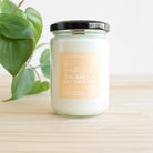 Candle in a glass jar with a label on a wooden surface with a plant in the background