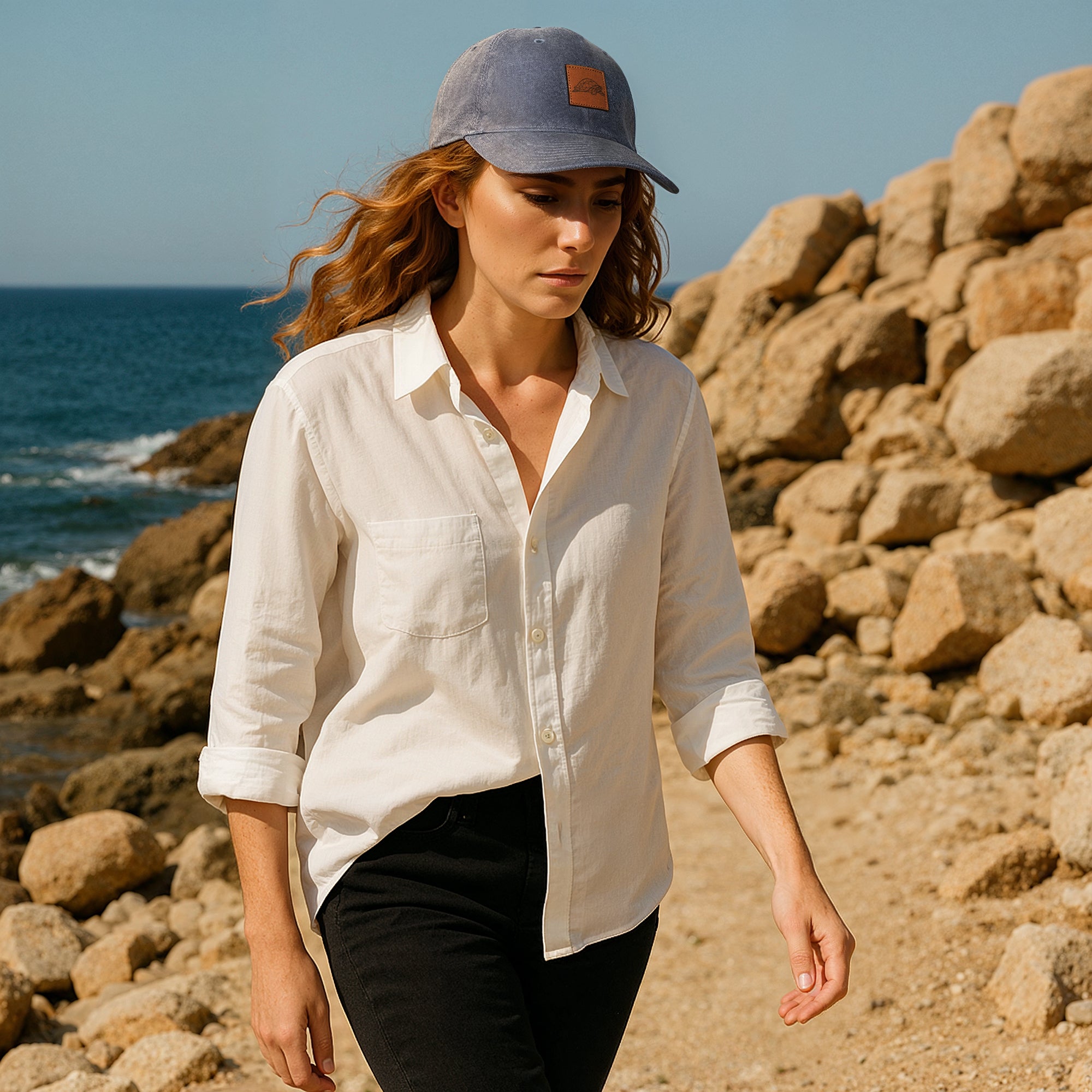 Woman wearing a white shirt and blue cap standing on rocky terrain by the ocean.