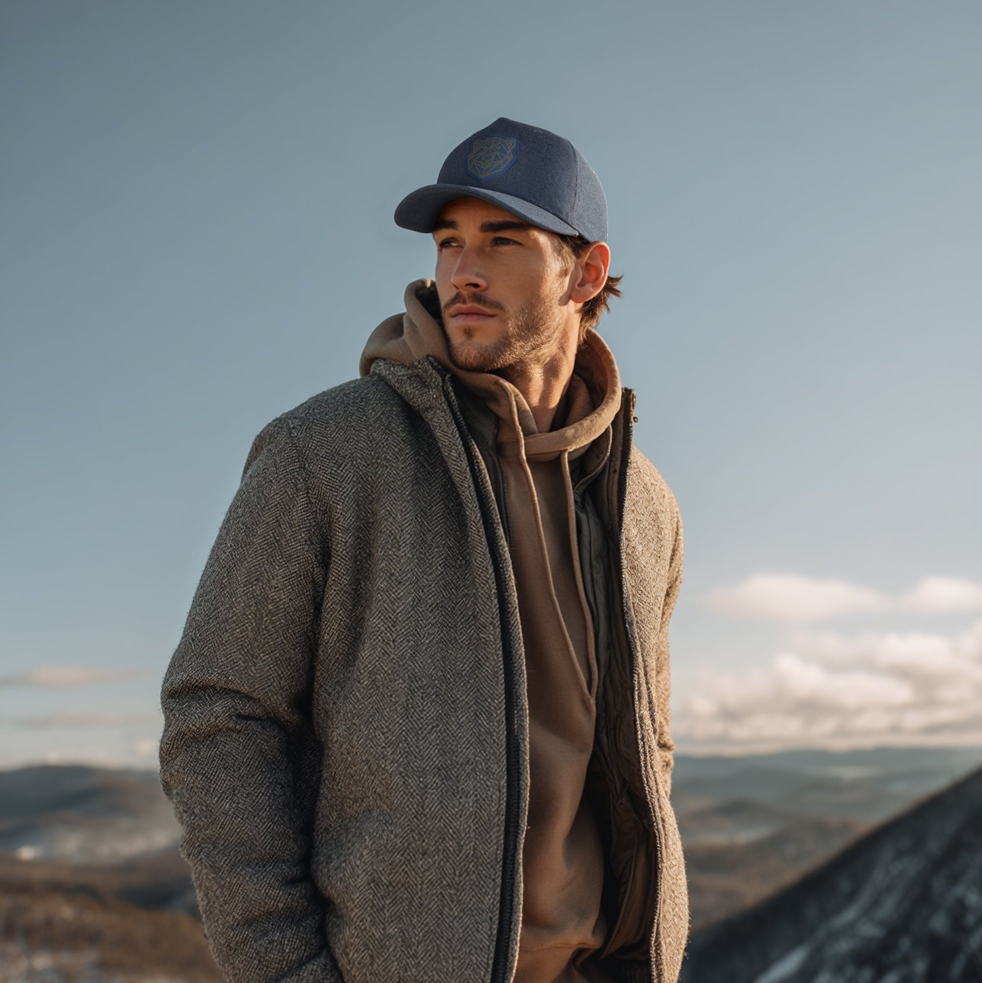 Man wearing a gray jacket and blue cap standing on a mountain with a clear sky.