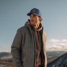 Man wearing a gray jacket and blue cap standing on a mountain with a clear sky.