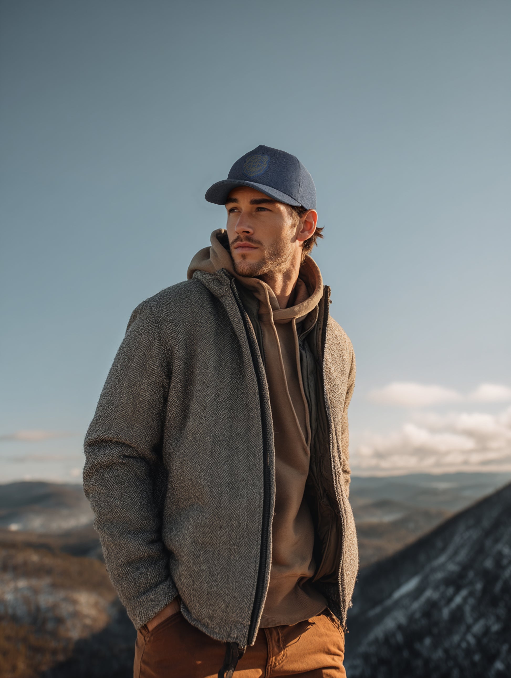 Man wearing a gray jacket and blue cap standing on a mountain with a clear sky.