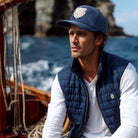 Man wearing a navy vest and cap on a boat with a scenic background