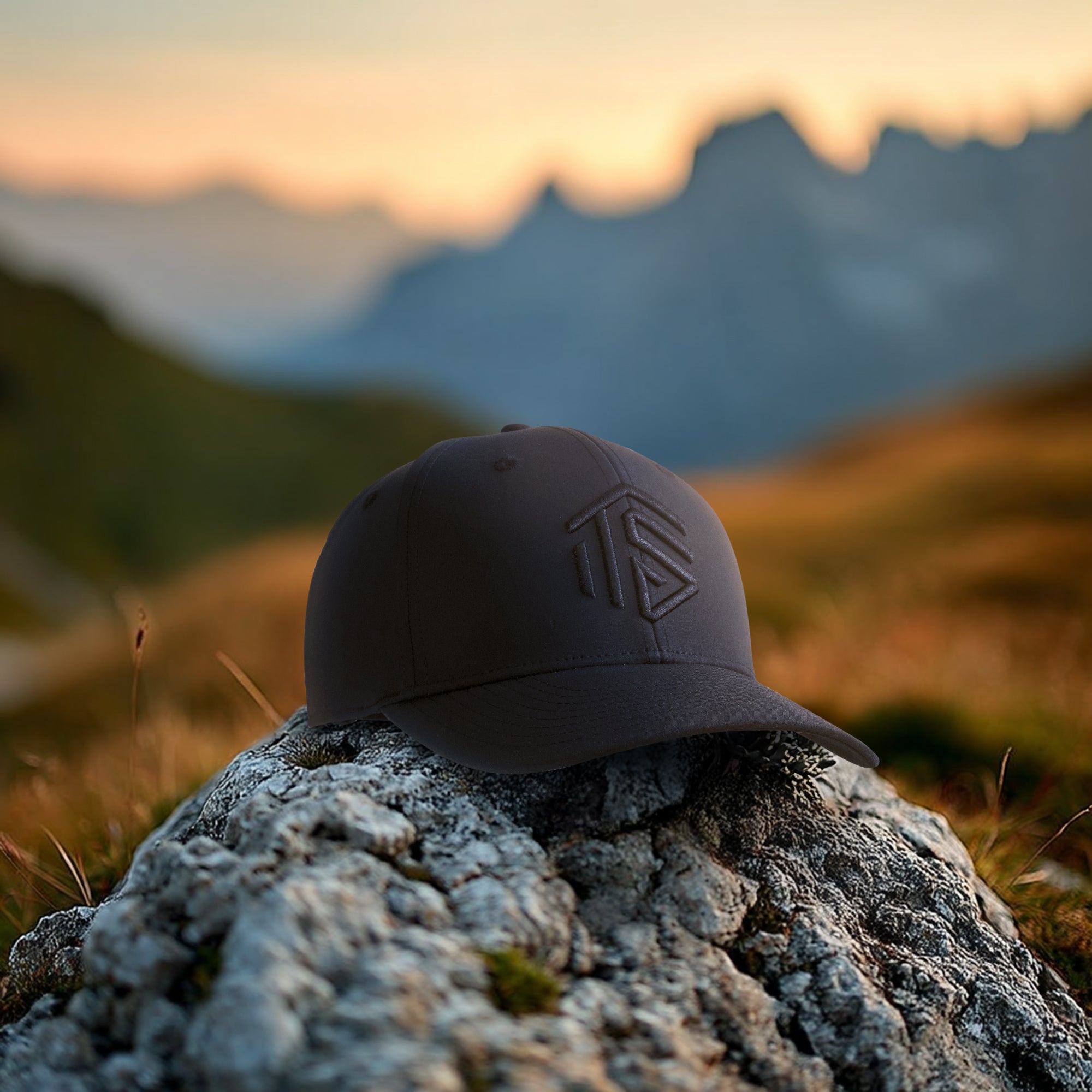 Dark-colored cap with a logo on a rock in a mountainous landscape during sunset.